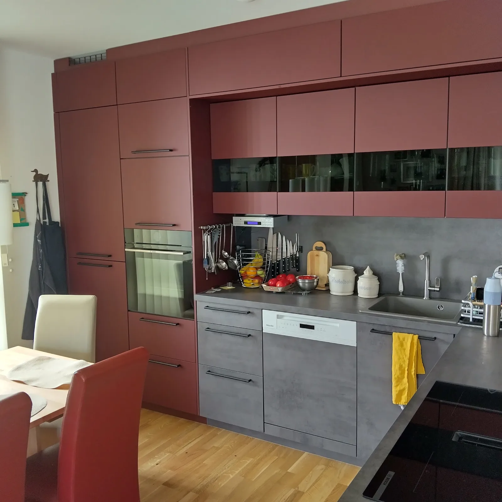 Eingebaute Küche mit weinroten Hochschränken, Glas-Oberschränken und Fronten in Betongrau, davor ein Esstisch